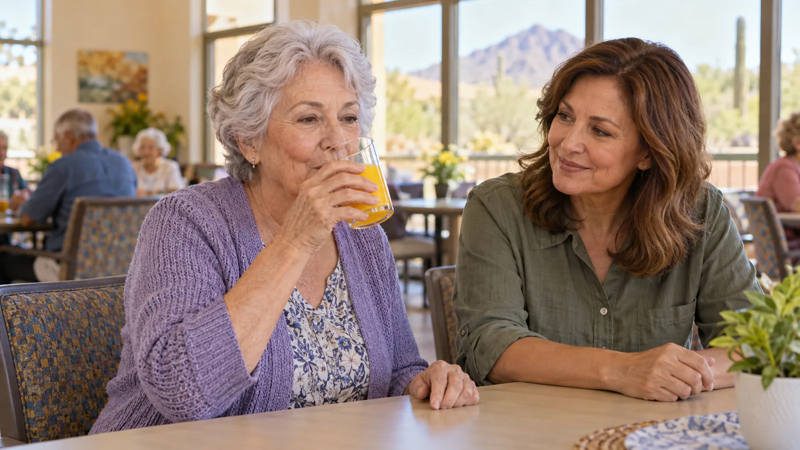An alert older woman drinks orange juice on her own while a helper sits beside her.
